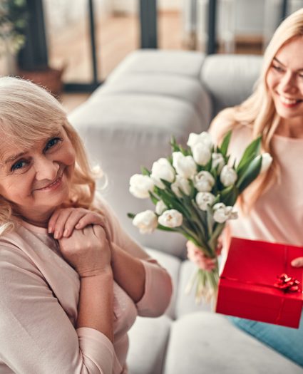 Senior woman and her attractive daughter spending time together at home and sitting on sofa. Happy Mothers' Day. Daughter giving present and flowers to her mom.